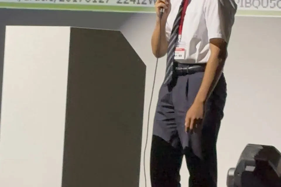 Toma on stage in a white shirt and tie, holding a handheld microphone beside a lectern, with the NOVA problem-statement slide projected behind him