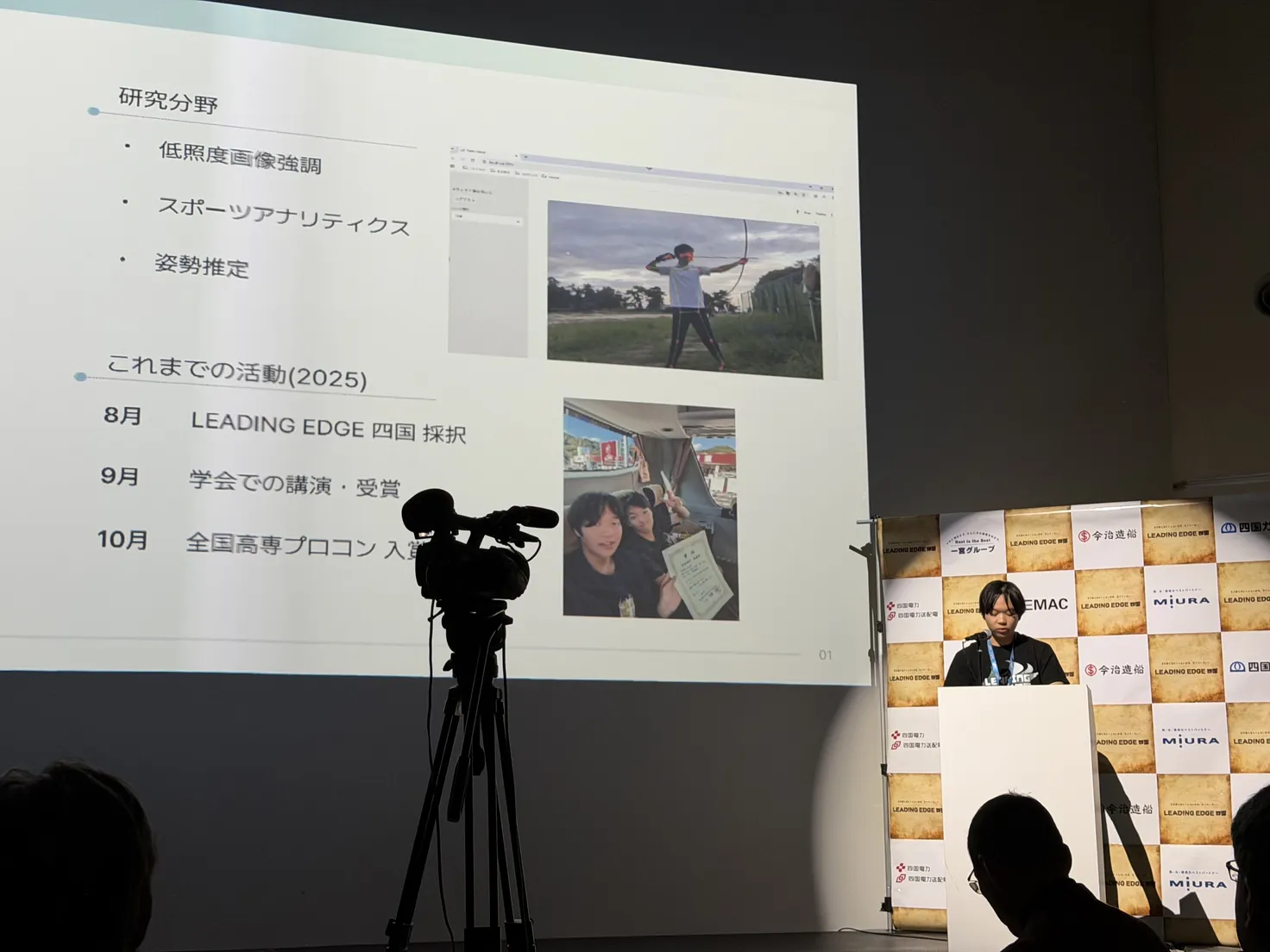 Toma Okugawa at a white podium at the LEADING EDGE Shikoku final report, beside a projected slide listing his research fields and 2025 activities; a video camera on a tripod stands in the foreground