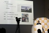 Toma Okugawa at a white podium at the LEADING EDGE Shikoku final report, beside a projected slide listing his research fields and 2025 activities; a video camera on a tripod stands in the foreground