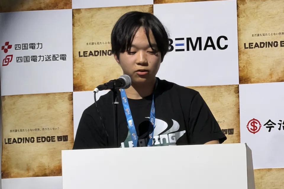 Toma speaking into a stand microphone at a white podium, wearing a black LEADING EDGE T-shirt and a lanyard, in front of a checkered backdrop of sponsor logos
