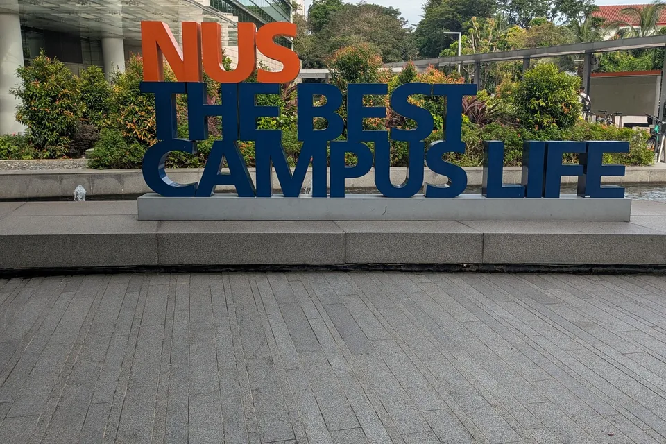 The NUS “The Best Campus Life” sign, orange and navy letters on a stone plinth in front of shrubs and a glass building