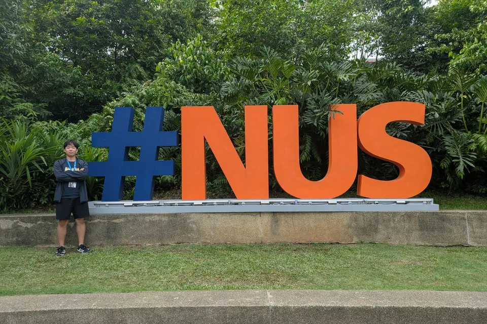 Toma standing with arms crossed beside the large blue-and-orange #NUS letters on the National University of Singapore campus, with tropical trees behind
