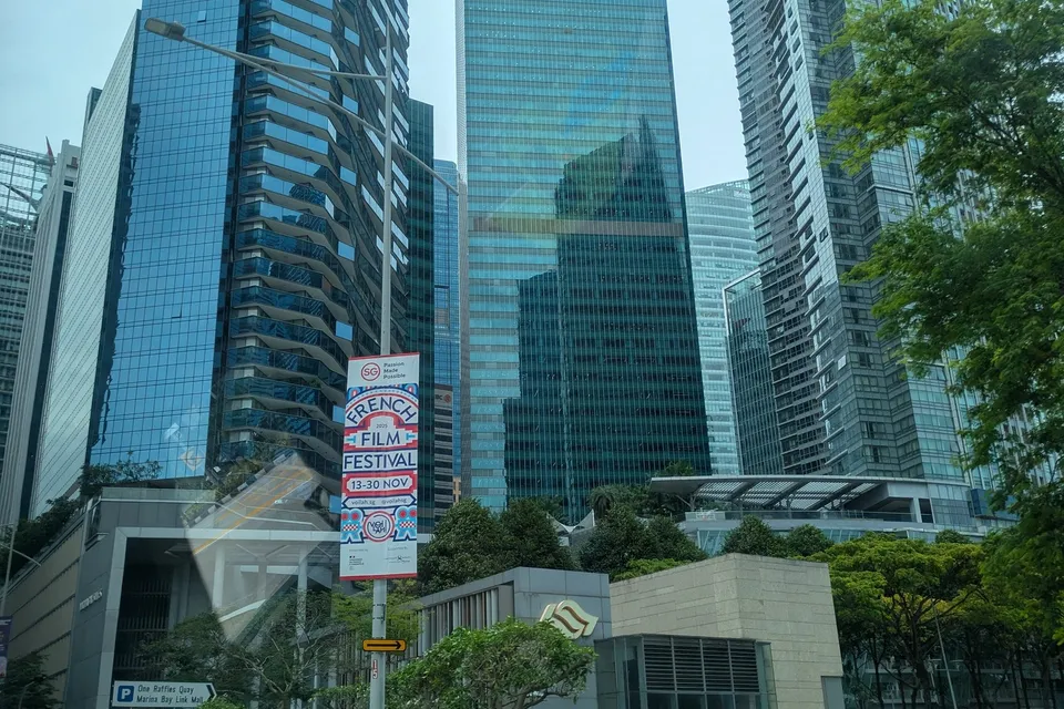 Glass office towers of Singapore’s Marina Bay district seen through a car window, with a French Film Festival banner dated November 2025 on a lamppost